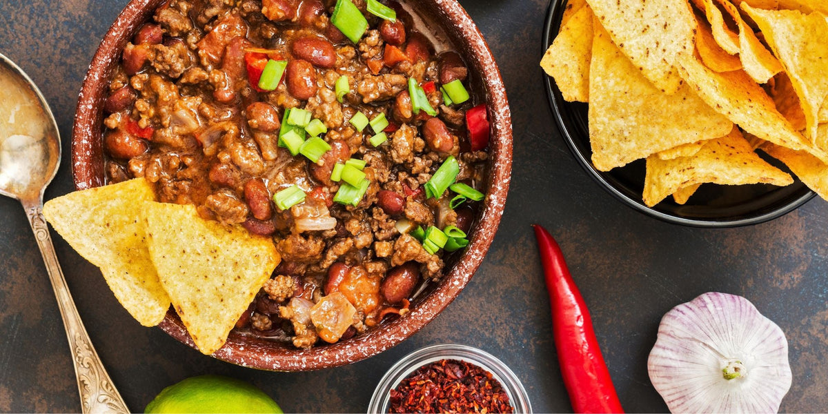 A bowl of chili with tortilla chips, a red chile pepper and a head of garlic on a dark table