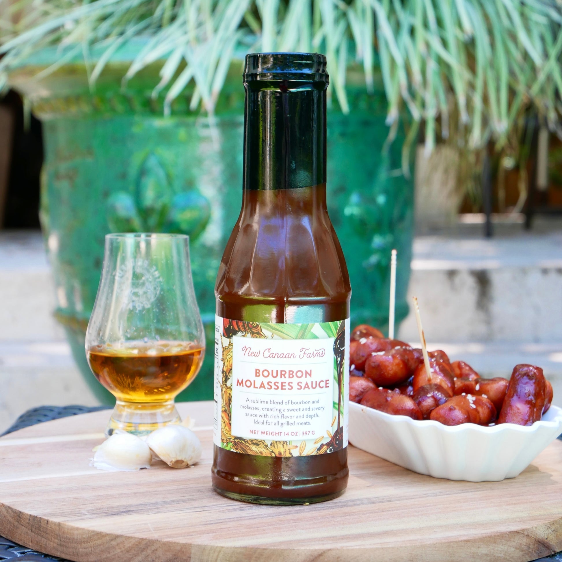 Bottle of New Canaan Farms  bourbon molasses sauce on a wooden board with a glass and bowl of grapes in the background.
