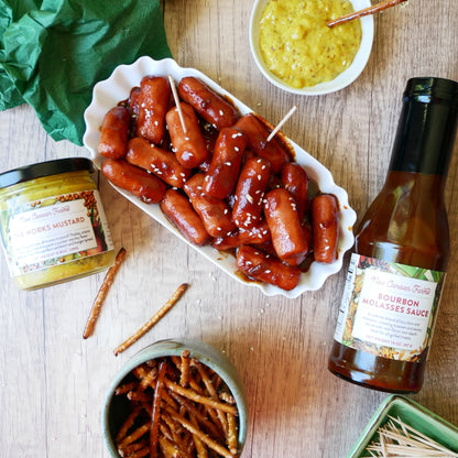 Platter of sausages with mustard, pretzels, and a bottle of New Canaan Farms Bourbon Molasses sauce on a wooden table.