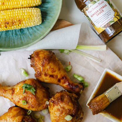 Baked chicken drumsticks with a plate of corn on the cob, ginger citrus sauce, and a brush on a white surface.