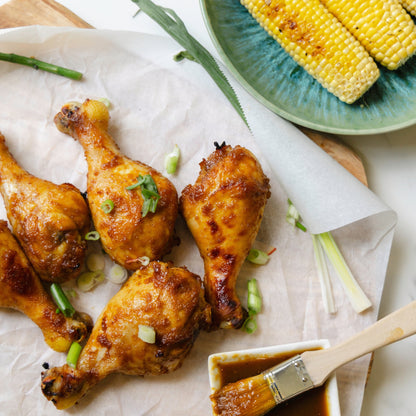 Baked chicken drumsticks with a brush and small bowl on parchment paper, corn on the cob in the background.
