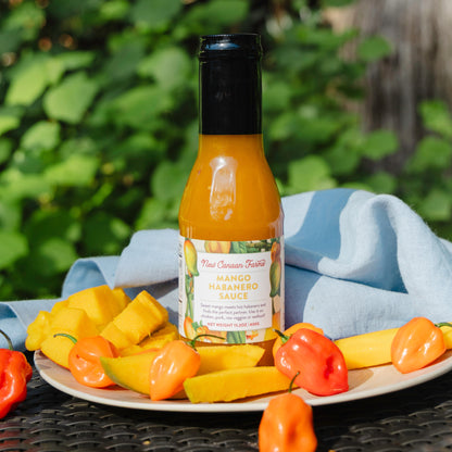 Bottle of New Canaan Farms Mango Habanero sauce on a plate with mangoes and habanero peppers outdoors.