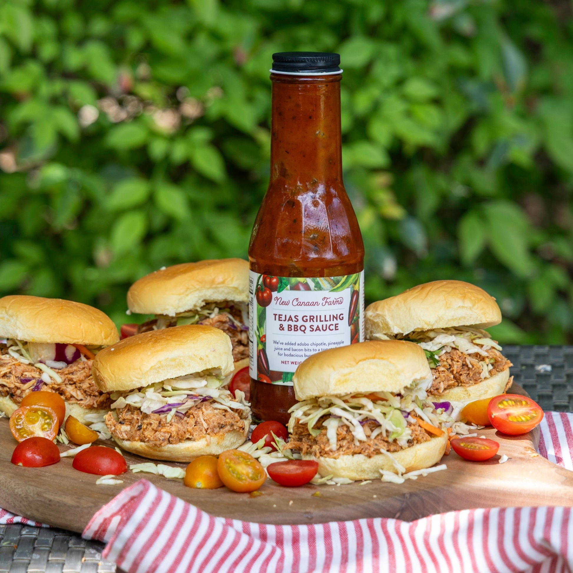 Burgers with New Canaan Farms Tejas Grilling & BBQ Sauce on a wooden board outdoors.
