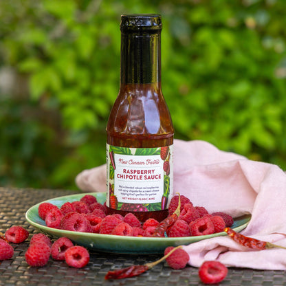 Bottle of New Canaan Farms Raspberry Chipotle Sauce on a plate with raspberries and a blurred green background