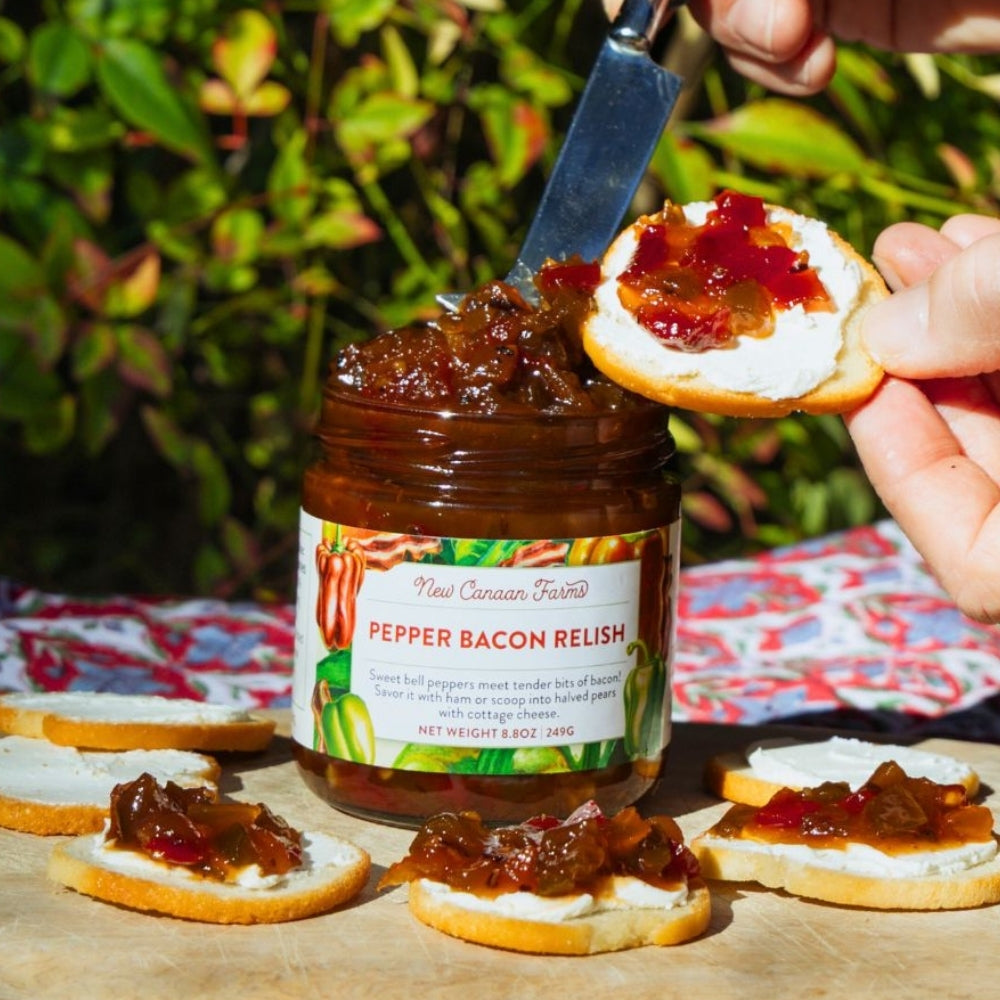 Jar of pepper bacon relish with crackers topped with relish on a table outdoors.
