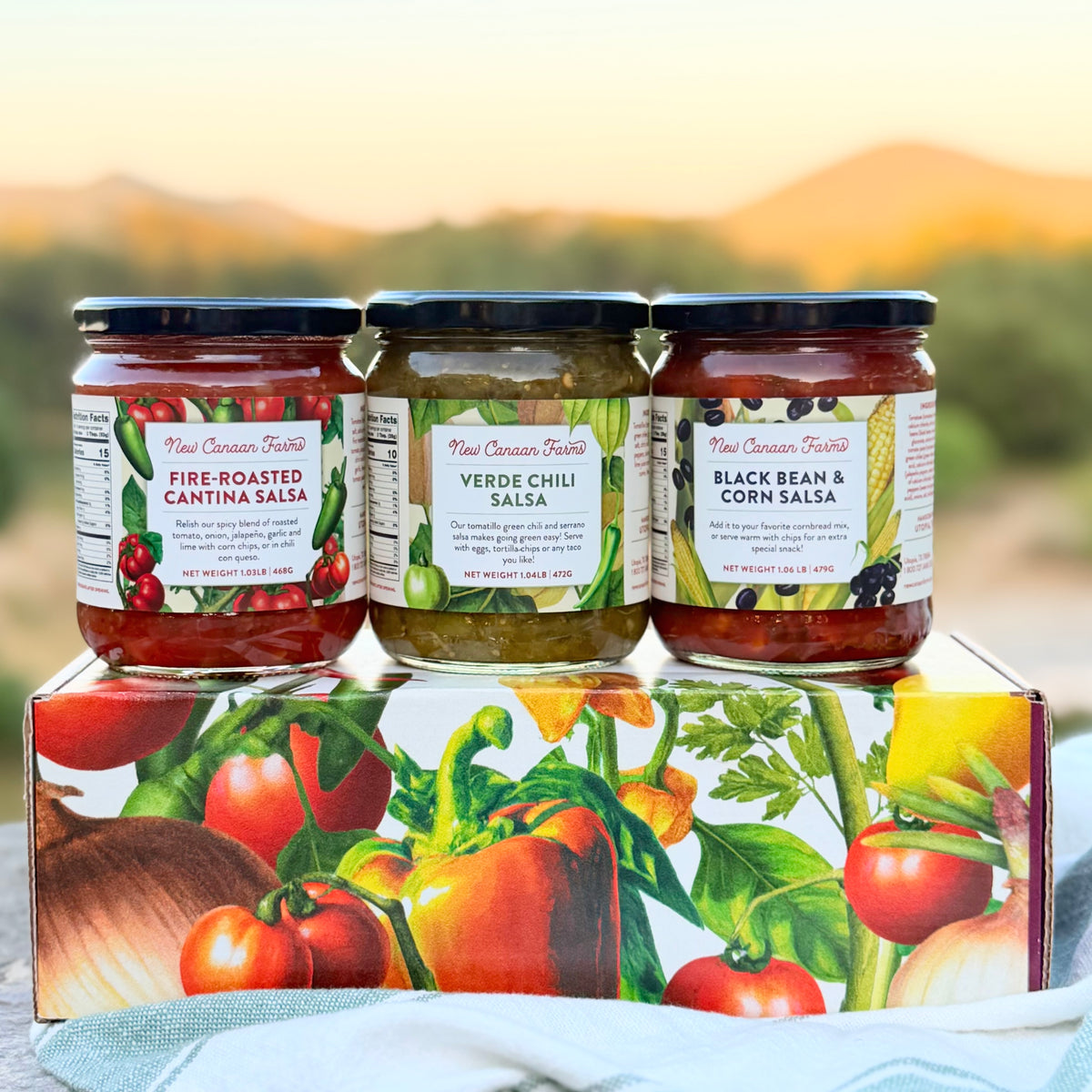 New Canaan Farms Fire-Roasted Cantina Salsa, Verde Chile Salsa and Black Bean & Corn Salsa jars on a vegetable-print box