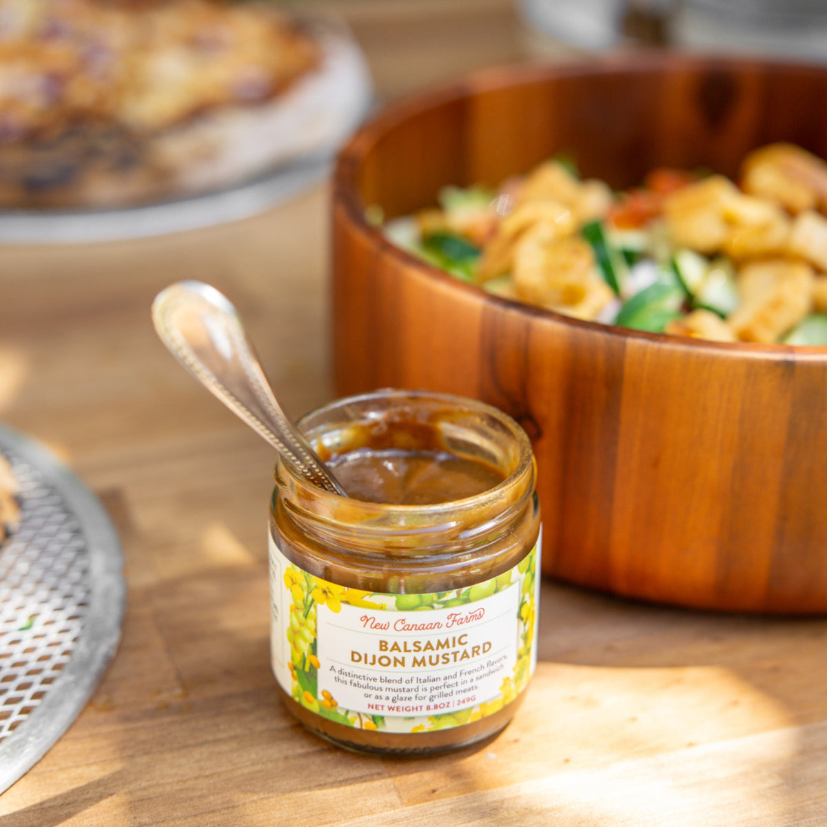 An open jar of New Canaan Farms Balsamic Dijon Mustard with a spoon, next to a wooden salad bowl