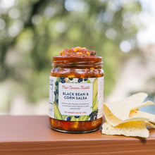 Jar of New Canaan Farms Black Bean & Corn Salsa with tortilla chips on a wooden surface outdoors.