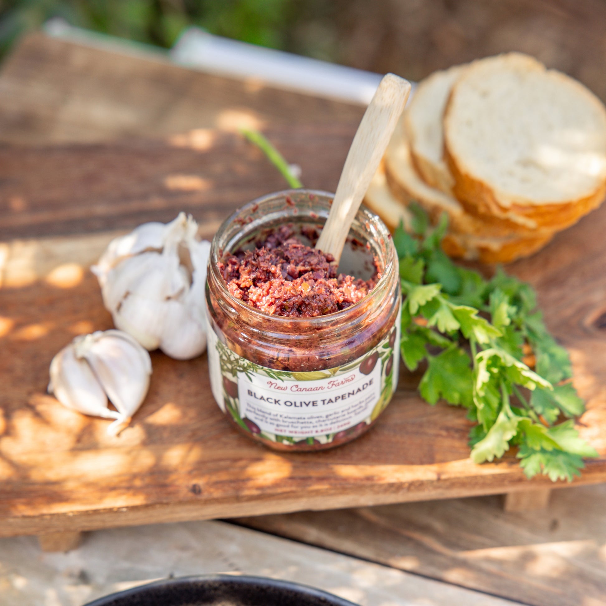 Jar of black olive tapenade with a spoon on a wooden board with bread and garlic.