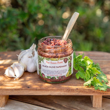Jar of black olive tapenade on a wooden surface with herbs and garlic, outdoor setting