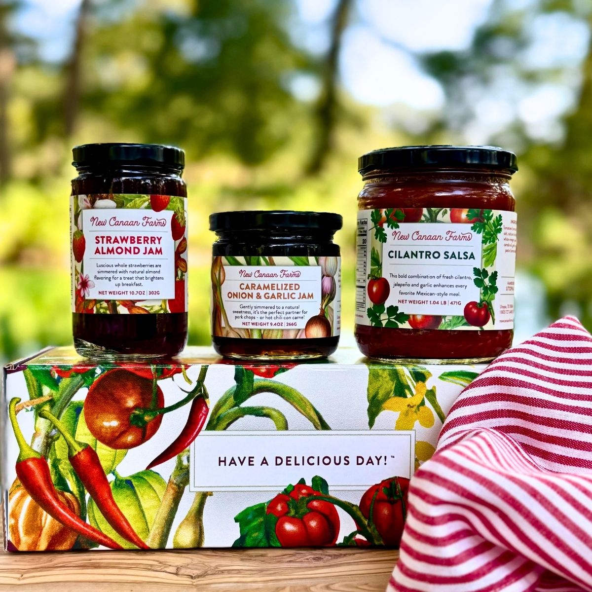 A jar of New Canaan Farms Strawberry Almond Jam, Caramelized Onion & Garlic Jam and Cilantro Salsa on a New Canaan Farms vegetable-inspired gift box, outside with a blurred background