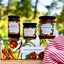 A jar of New Canaan Farms Strawberry Almond Jam, Caramelized Onion & Garlic Jam and Cilantro Salsa on a New Canaan Farms vegetable-inspired gift box, outside with a blurred background