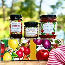 A jar of New Canaan Farms Strawberry Almond Jam, Caramelized Onion & Garlic Jam and Cilantro Salsa on a New Canaan Farms holiday gift box, outside with a blurred background