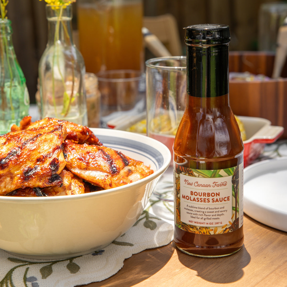 A bottle of New Canaan Farms Bourbon Molasses Sauce beside a bowl of grilled chicken on an outdoor table