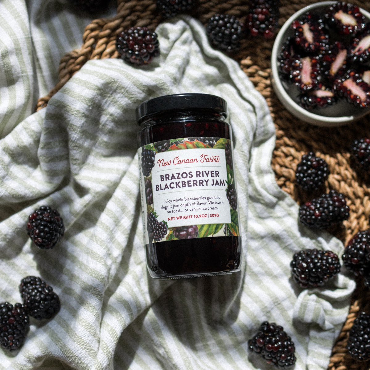 New Canaan Farms Brazos River Blackberry Jam lying on a striped towel, surrounded by blackberries