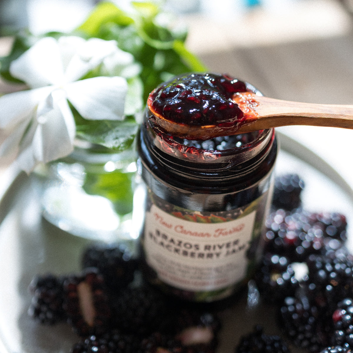 Close-up of a wooden spoon of Brazos River Blackberry Jam on the open jar, with blackberries and a white flower