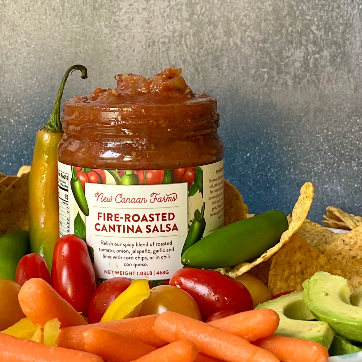 New Canaan Farms Fire-Roasted Cantina Salsa jar with tortilla chips, jalapeños, cherry tomatoes, baby carrots and avocado