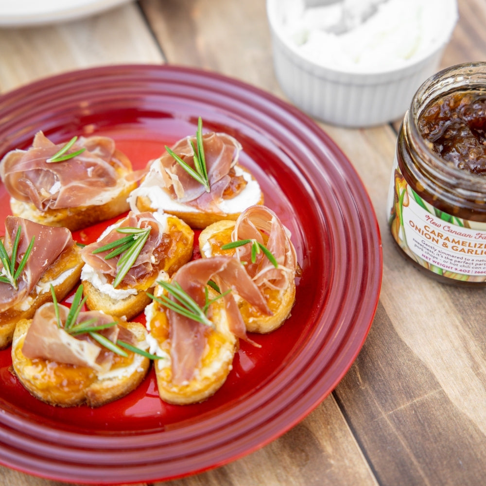 Red plate with small sandwiches topped with prosciutto and arugula, next to a jar of spread on a wooden table.