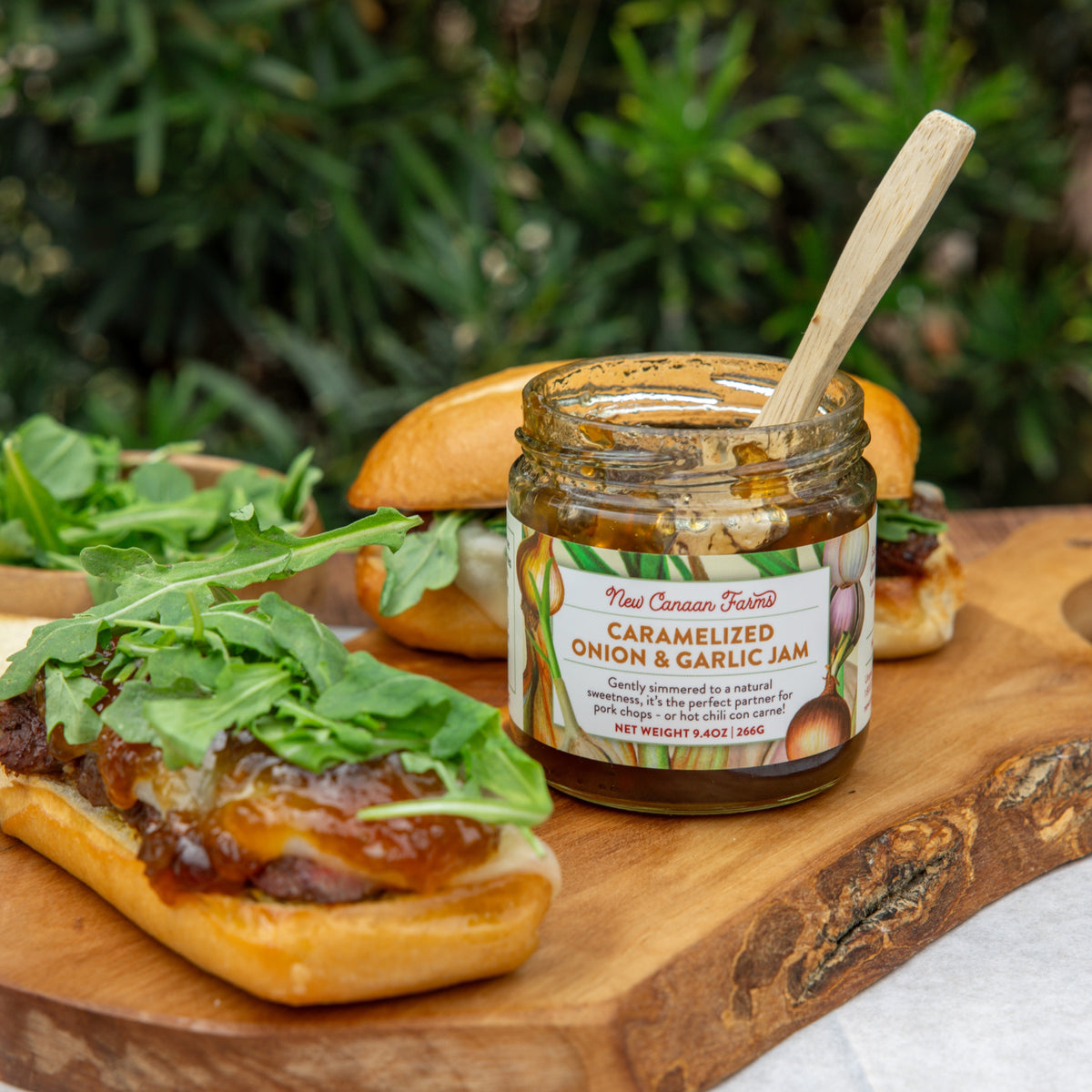 Jar of caramelized onion and garlic jam on a wooden board with burgers and arugula.