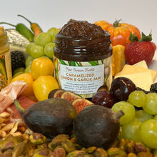 Jar of caramelized onion and garlic jam surrounded by fruits, vegetables, and cheese on a white background