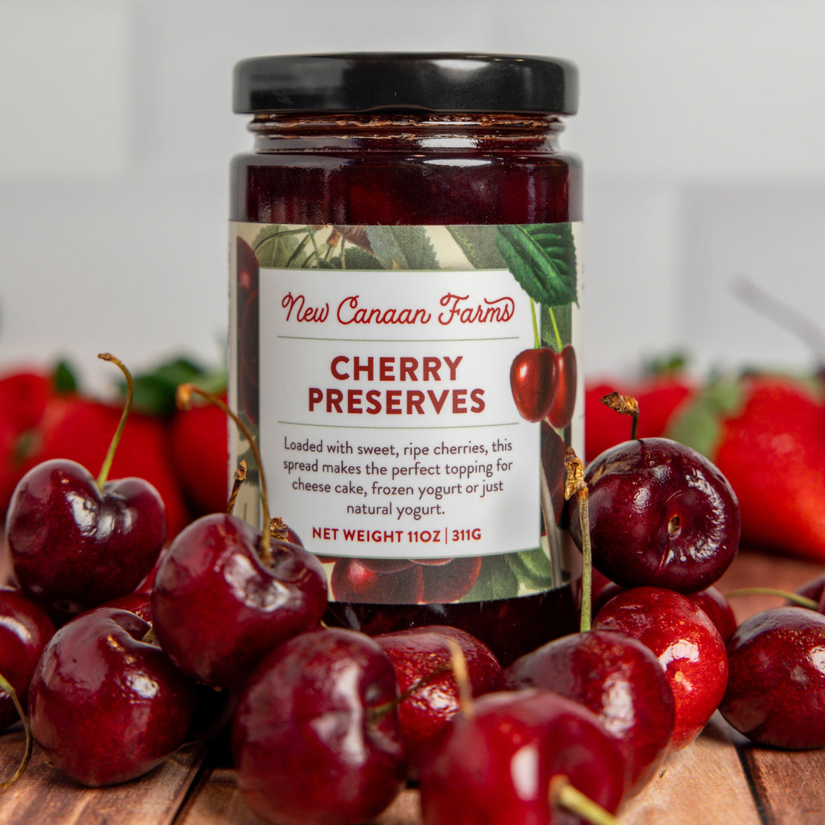 Jar of New Canaan Farms Cherry Preserves surrounded by cherries on a wooden surface.
