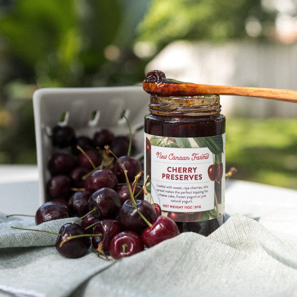Jar of New Canaan Farms Cherry Preserves with a wooden spoon and cherries on a clothed surface.