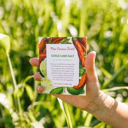 Hand holding a New Canaan Farms Chile Lime Salt packet with a green field background