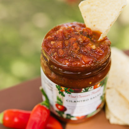 Jar of cilantro salsa with a tortilla chip on top, surrounded by red peppers and tortilla strips.