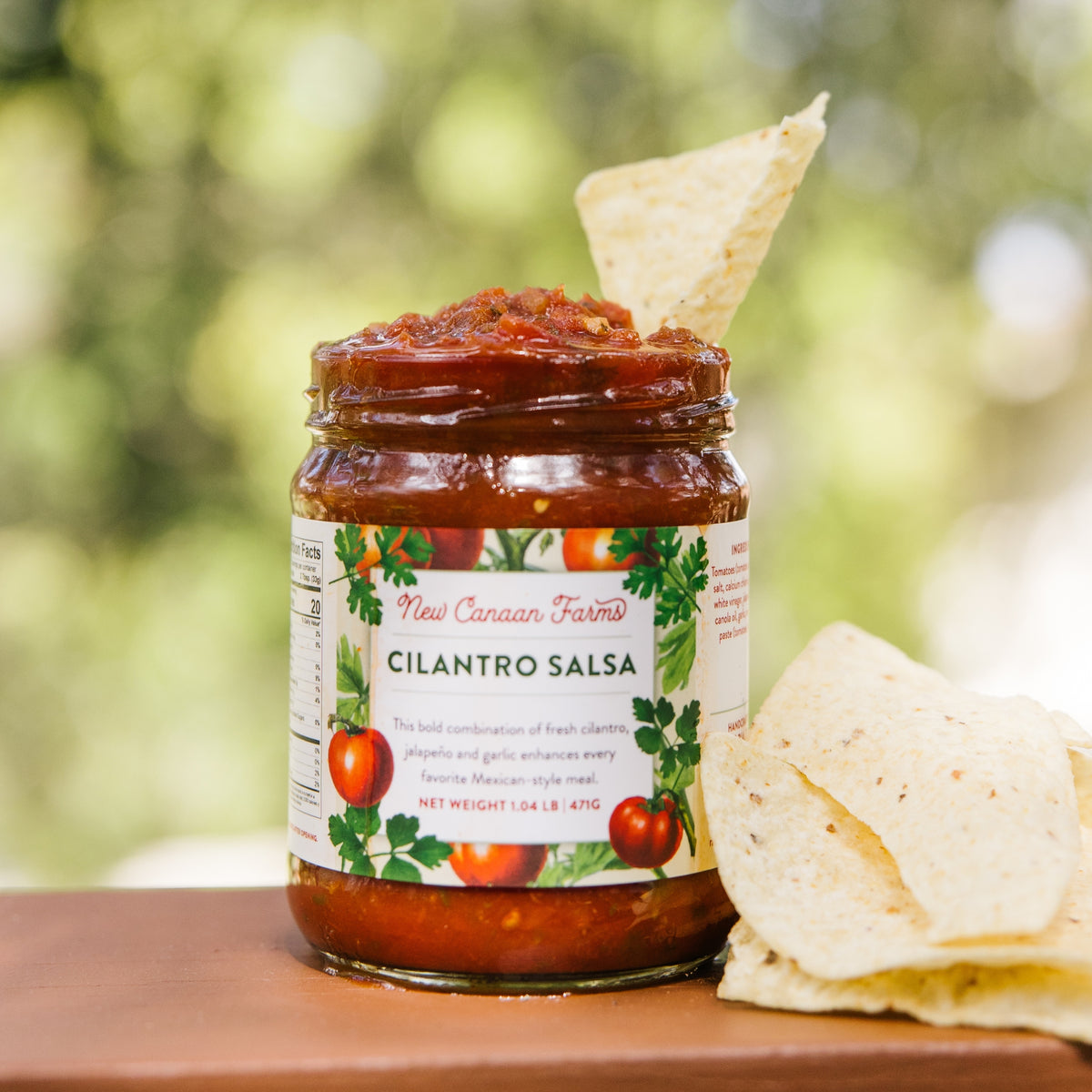 Jar of cilantro salsa with a tortilla chip on top, set against a blurred green outdoor background.