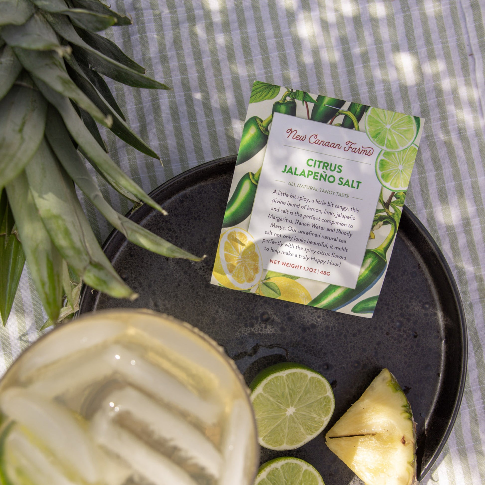Citrus Jalapeno Salt package on a black plate with a pineapple and lime slices, on a striped fabric background.