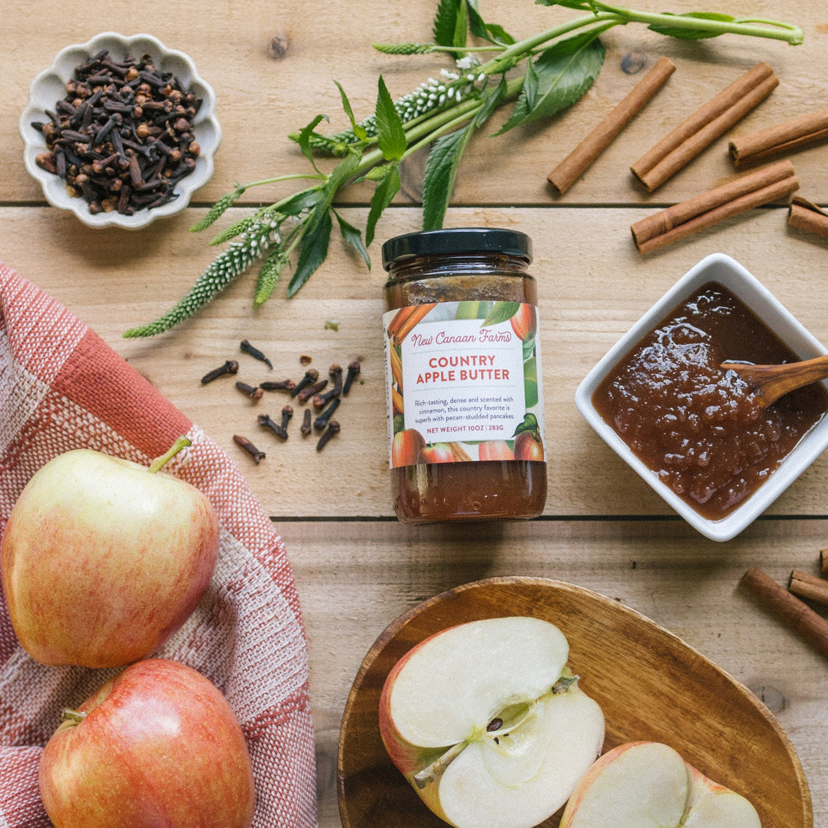 New Canaan Farms Country Apple Butter jar on a wooden table with a bowl of apple butter, apples and cinnamon