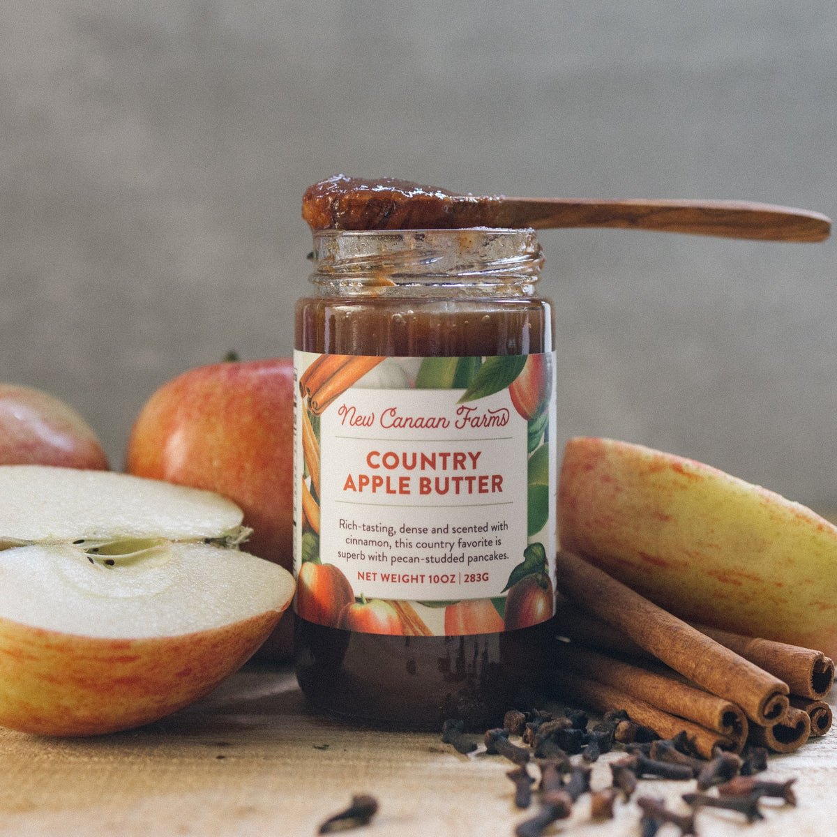 New Canaan Farms Country Apple Butter jar with a spoonful on top, beside apples, cinnamon sticks and cloves