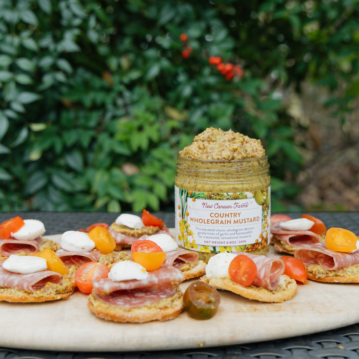 Jar of New Canaan Farms country wholegrain mustard on a platter with crackers and toppings outdoors.