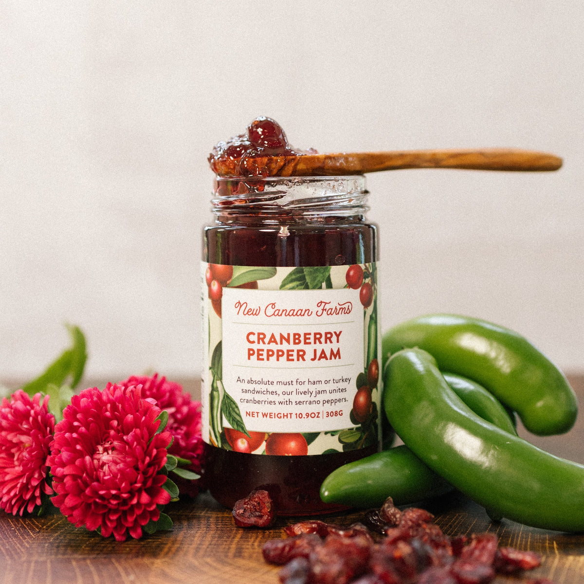 New Canaan Farms Cranberry Pepper Jam jar with a spoonful on top, beside serrano peppers and dried cranberries
