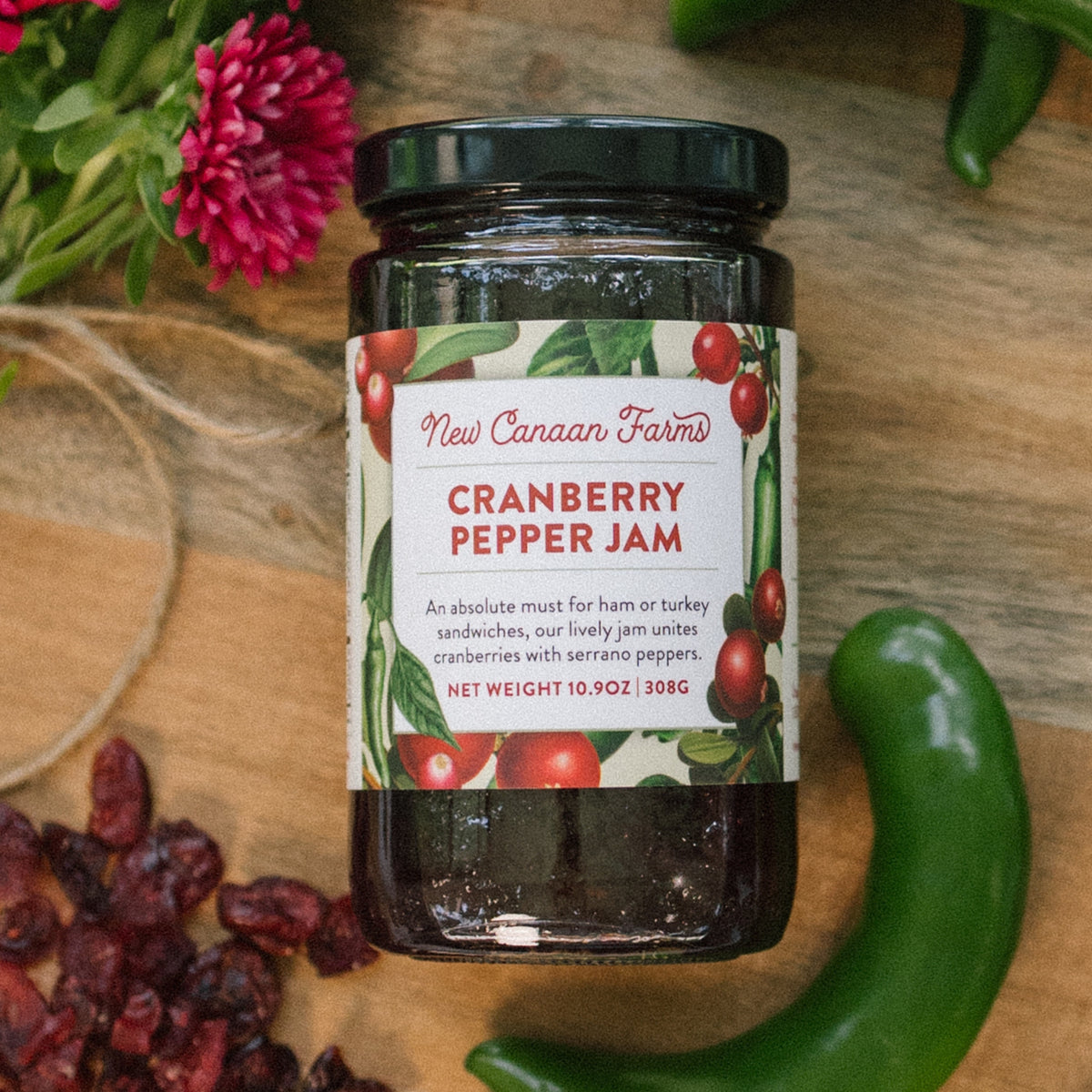 New Canaan Farms Cranberry Pepper Jam jar on a wooden board with dried cranberries and a serrano pepper