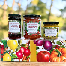 Jars of New Canaan Farms Cherry Preserves, Fire-Roasted Cantina Salsa and Olive Rosemary Dijon Mustard displayed on top of a colorful fruit and vegetable gift box outdoors.