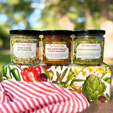 New Canaan Farms Three Cheese Artichoke Dip, Sun-Dried Tomato & Basil Mustard and Green Olive Tapenade sitting on a gift box, outside in front of trees