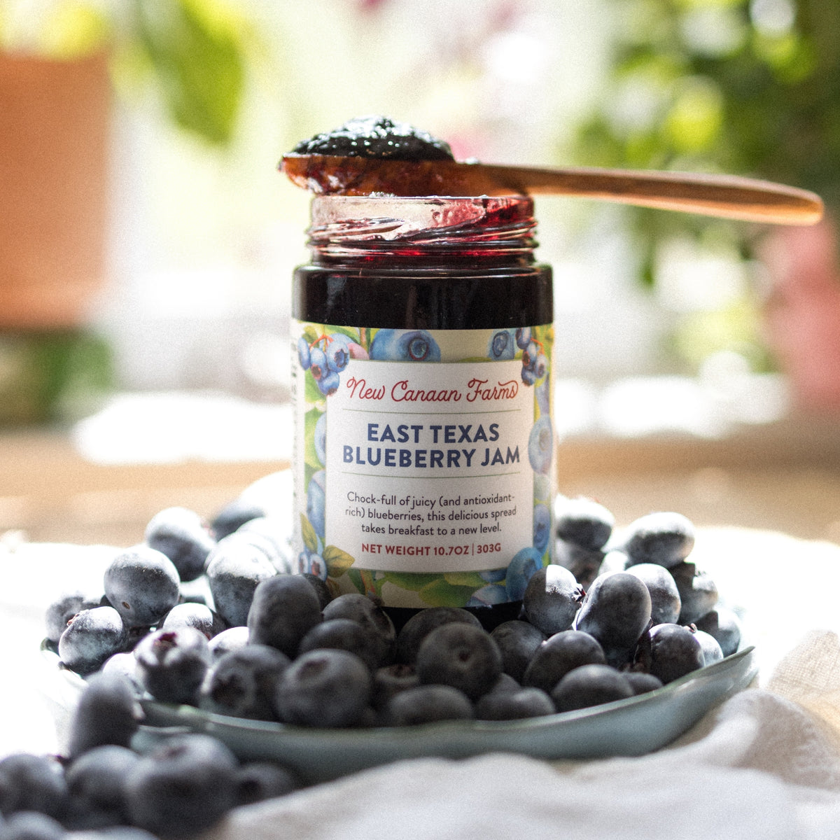 Jar of New Canaan Farms East Texas Blueberry Jam with a spoonful on top, sitting in a plate of fresh blueberries