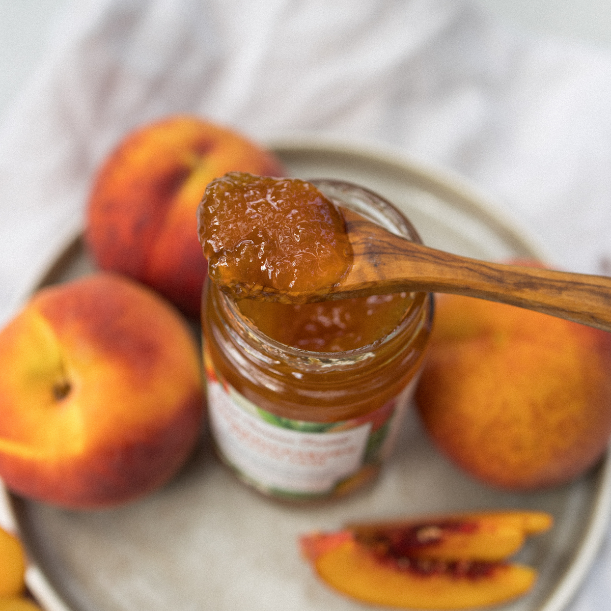 Wooden spoon of Fredericksburg Peach Jam over the open jar, with fresh peaches behind