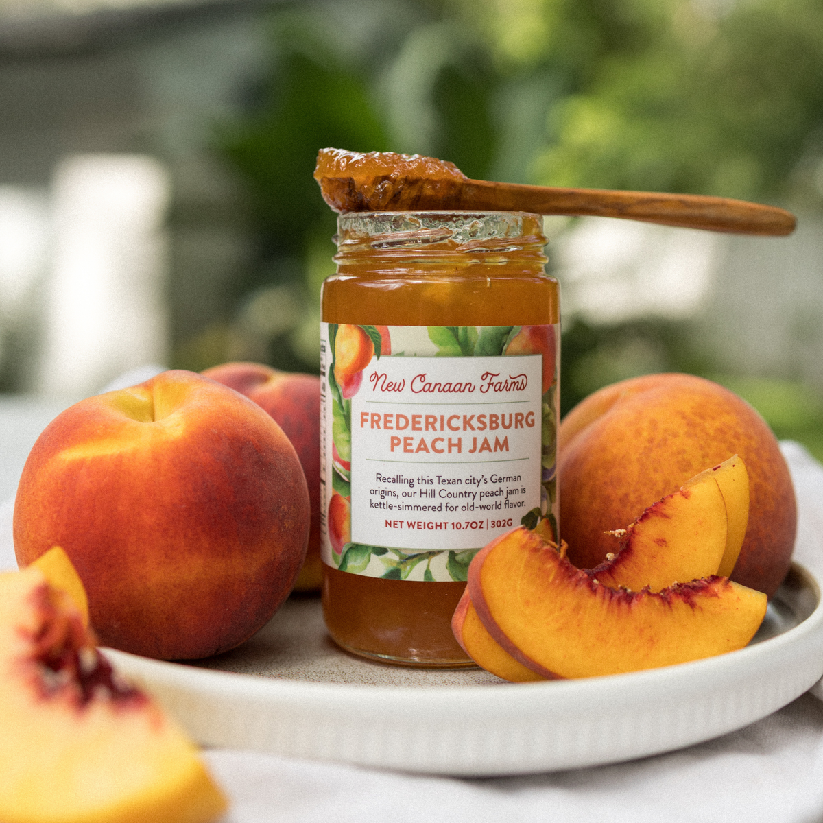 A jar of New Canaan Farms Fredericksburg Peach Jam, photographed on a white plate outside, surrounded by fresh peaches