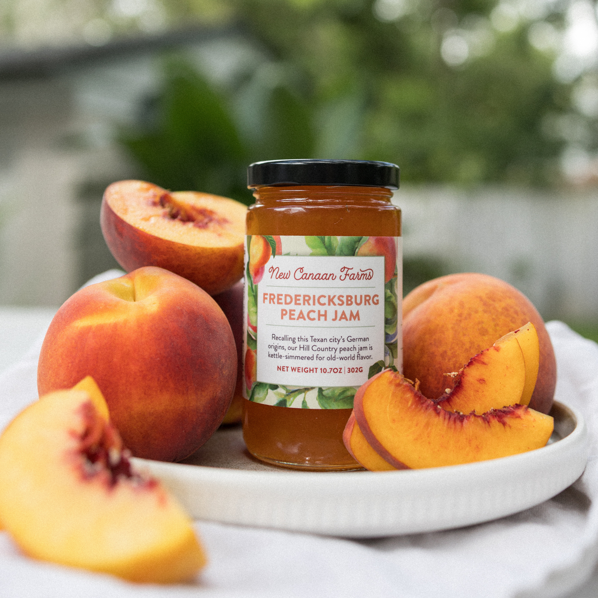 Jar of New Canaan Farms Fredericksburg Peach Jam on a plate with whole and sliced fresh peaches