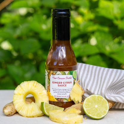 A jar of New Canaan Farms Ginger Citrus sauce, photographed outside on a white cloth, surrounded by fresh slices of pineapple and limes