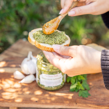 Person spreading green olive tapenade on a slice of bread with a wooden spoon and garlic cloves on a wooden surface.