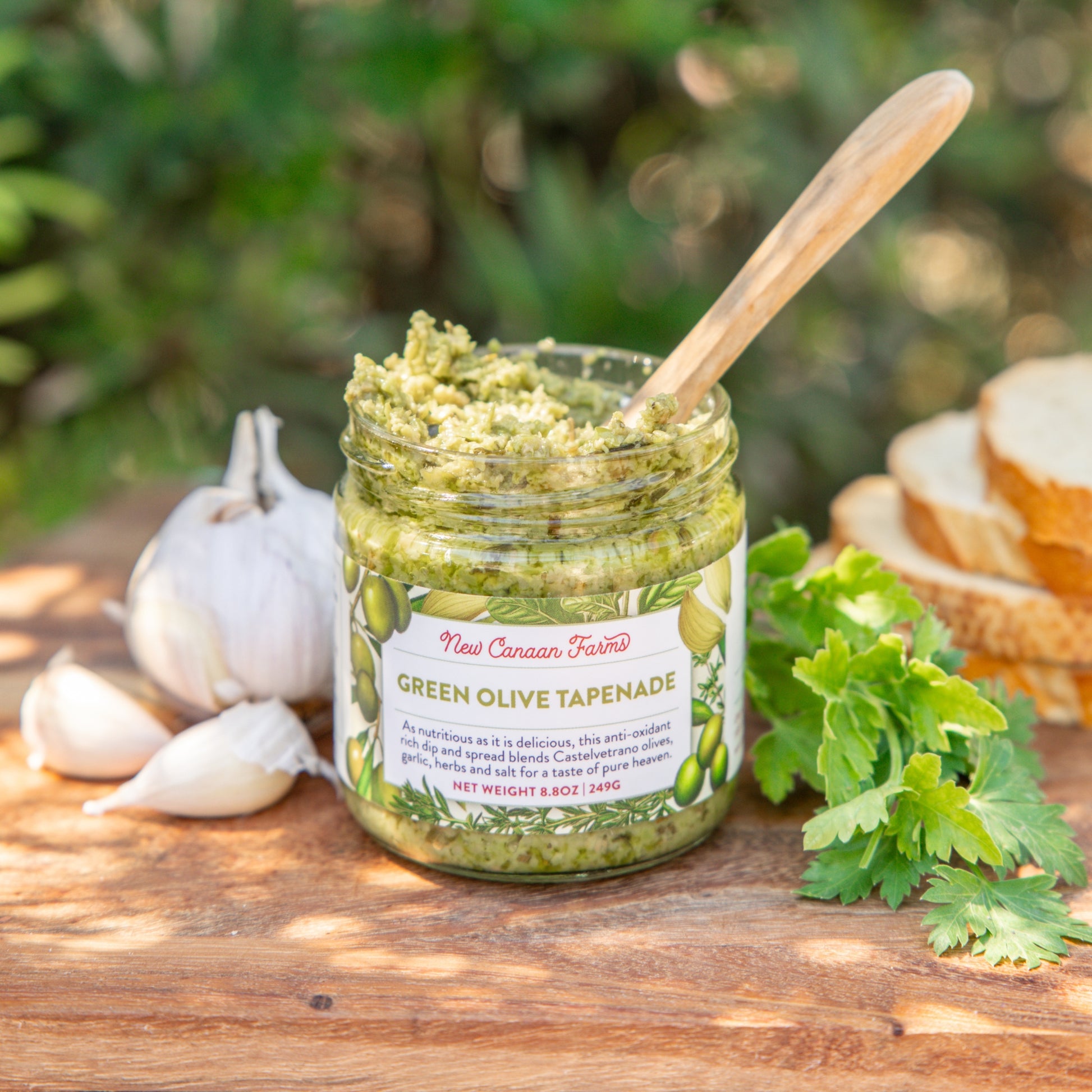 Jar of green olive tapenade with a wooden spoon on a wooden surface with garlic and bread in the background.
