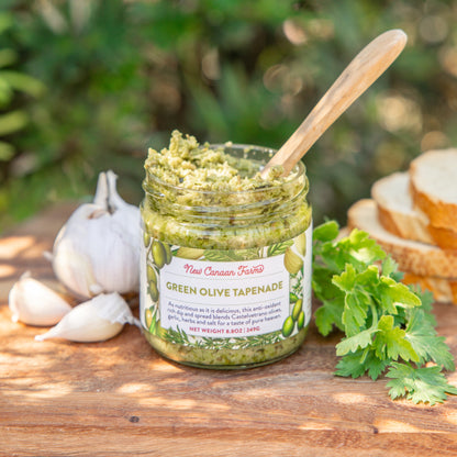 Jar of green olive tapenade with a wooden spoon on a wooden surface with garlic and bread in the background.