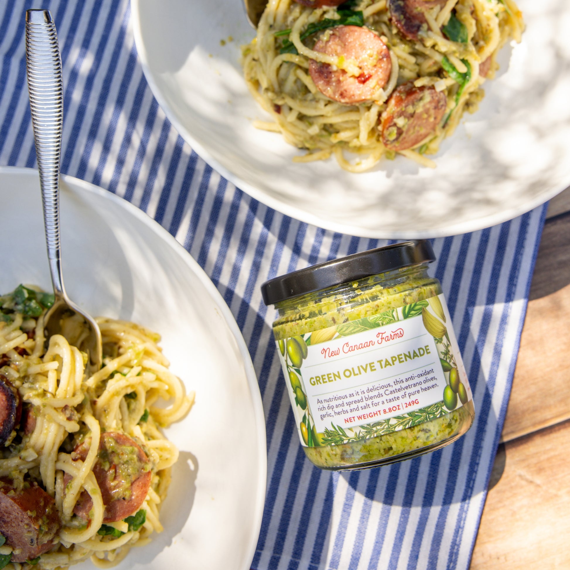 Two plates of pasta with a jar of green olive tapenade on a striped tablecloth.