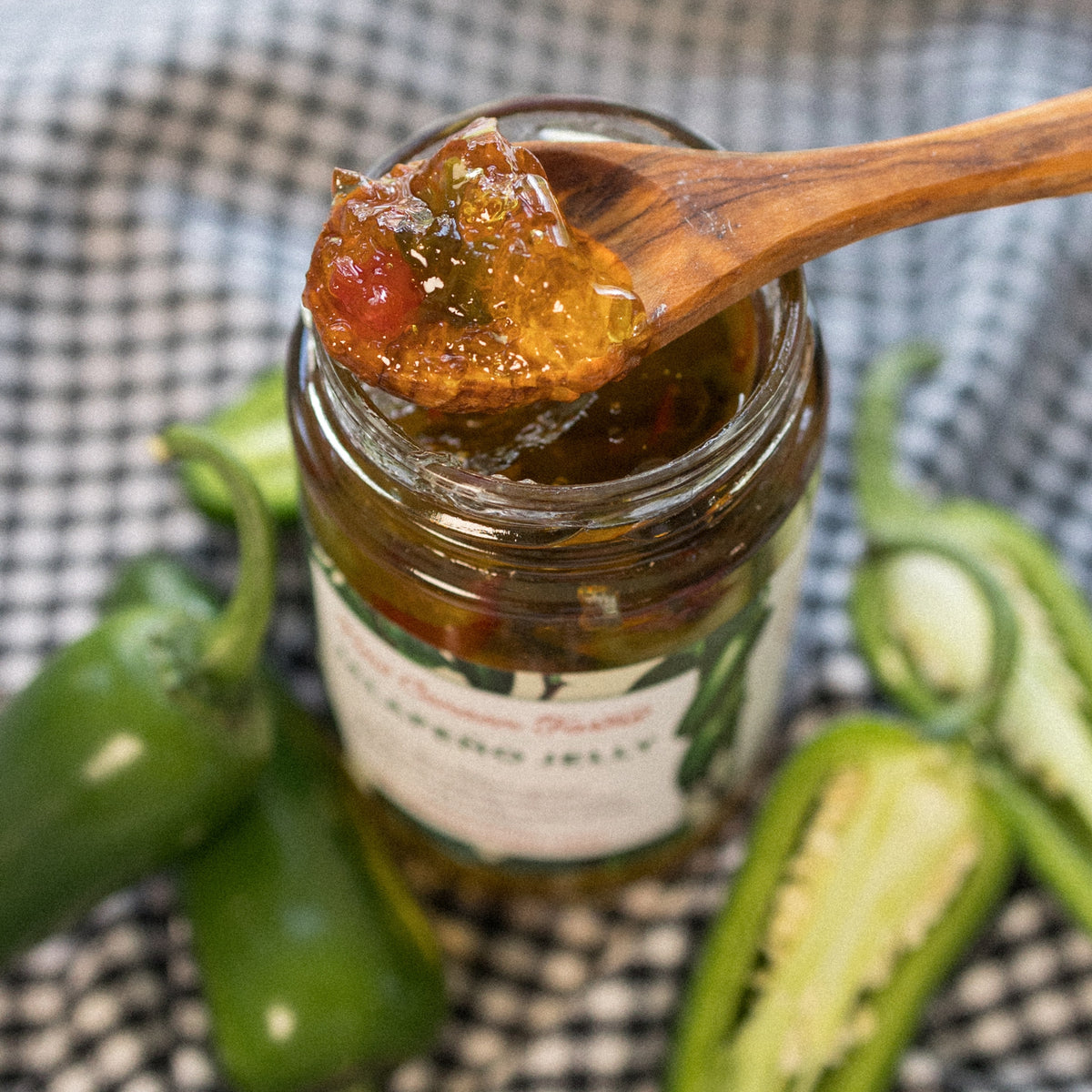 Wooden spoon of Jalapeño Jelly with red pepper pieces over the open jar, with fresh jalapeños nearby