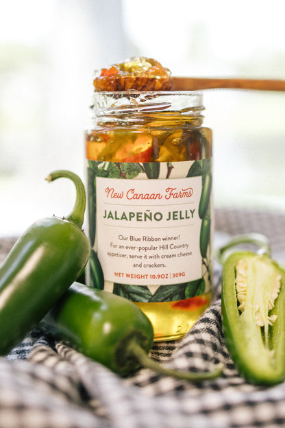 A jar of New Canaan Farms Jalapeño Jelly with a wooden spoon spooning out some of the contents. The jar has some fresh jalapeños around it