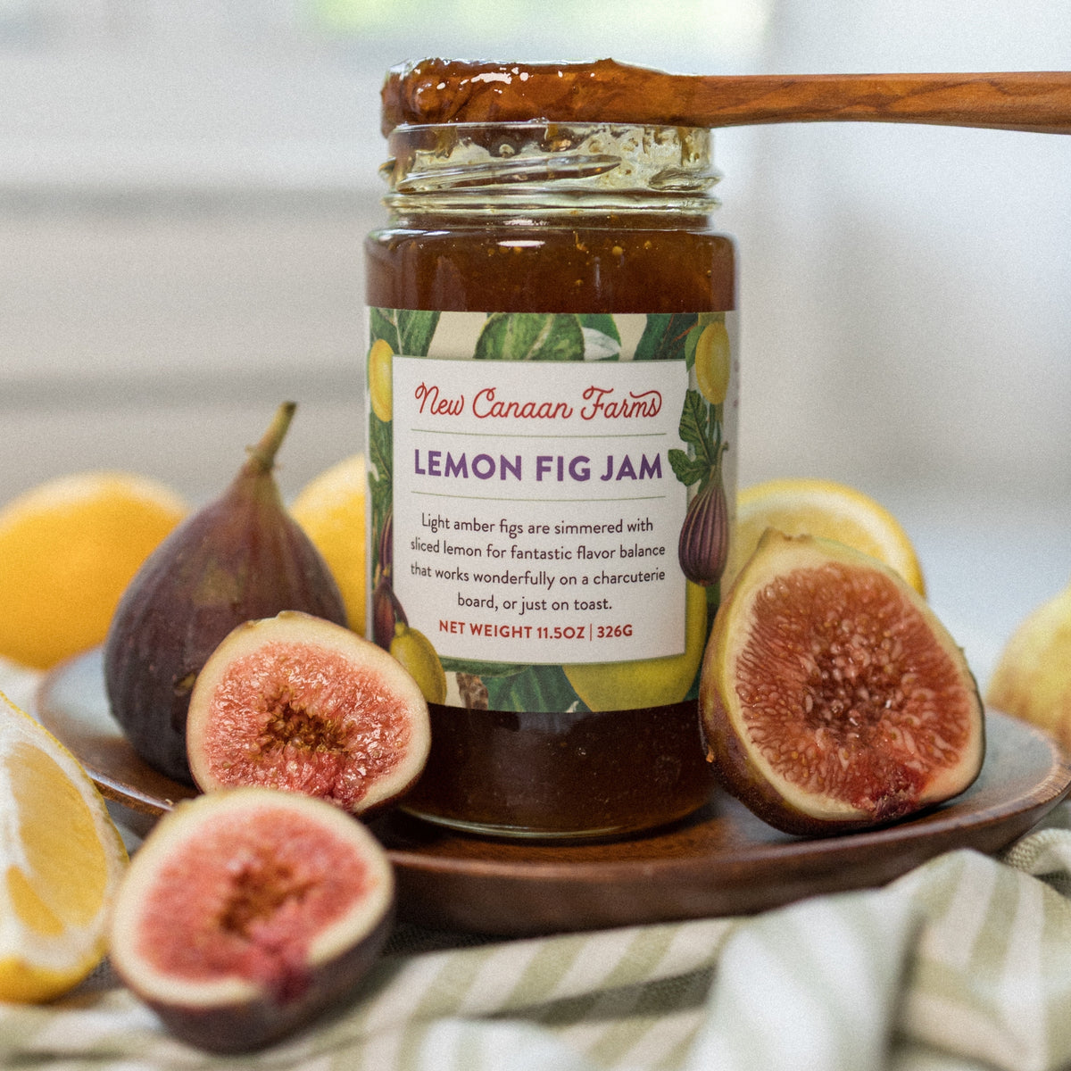 A jar of New Canaan Farms Lemon Fig Jam, on a wooden plate, surrounded by fresh figs and lemons