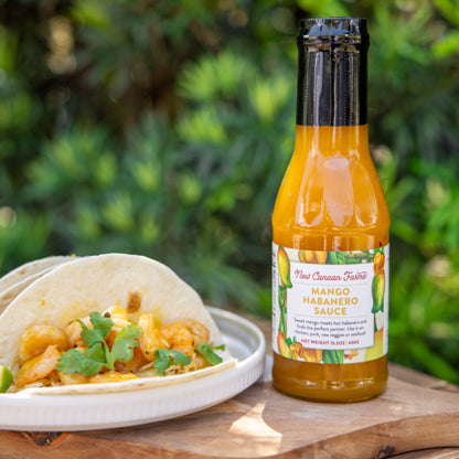 Bottle of Mango Habanero sauce next to tacos on a wooden surface with a green outdoor background
